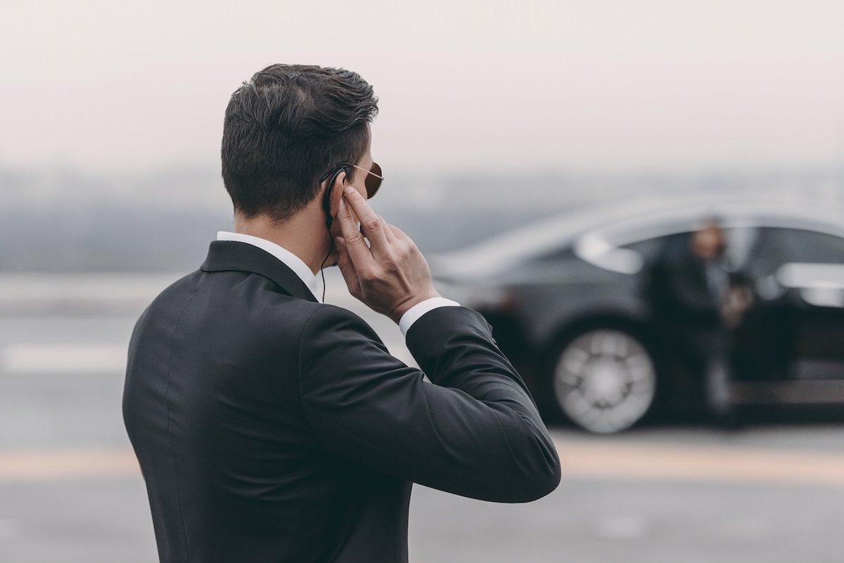 Security officer in a dark suit with an earpiece standing beside an executive car